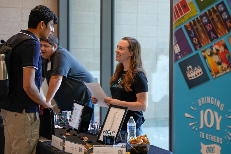 A woman stands behind a recruitment booth, talking with a student. She is holding the student’s resume.