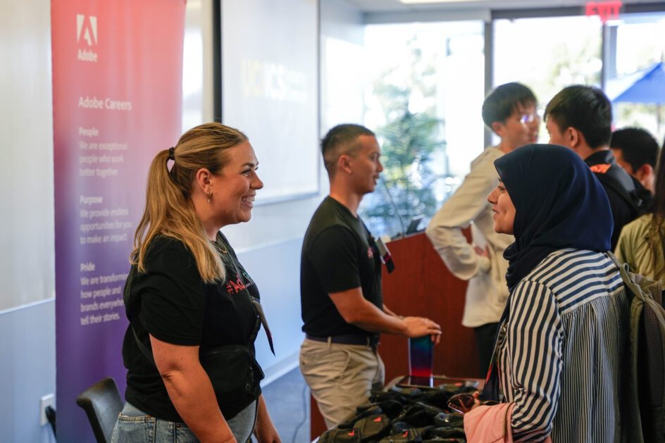 A woman stands behind a table, speaking a with a female Ph.D. student. Another recruiter speaks with other students in the background.