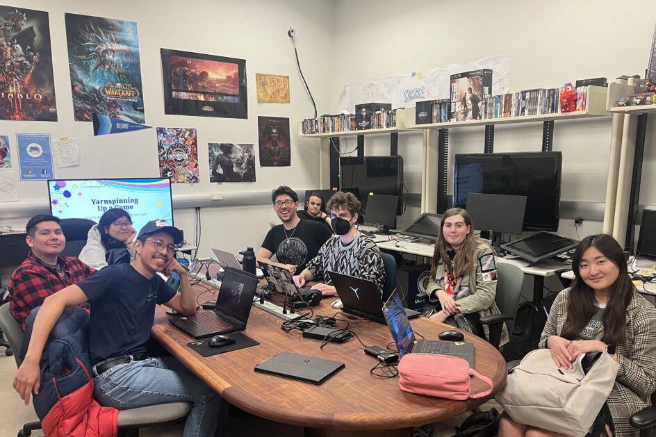 Eight students sitting around a table with laptops in a room with video games and posters.